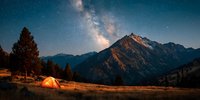 Illuminated camping tent in mountain valley under starry night sky with Milky Way galaxy arching over rugged alpine peaks.