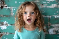 Excited young Caucasian girl with curly blonde hair and bright blue eyes showing surprised expression against weathered turquoise wooden wall background.