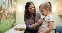Female pediatrician examining young blonde girl with stethoscope in colorful medical office. Healthcare professional providing checkup for child patient.