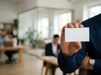 Businessman holding blank white business card in modern office environment with colleagues working in background.