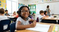 Young African American girl with glasses and braids smiling at desk in elementary school classroom with diverse students and teacher at whiteboard.