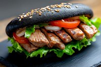 Grilled beef slices with fresh vegetables and lettuce in black charcoal burger bun on dark slate background, close up view of gourmet sandwich.