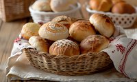 Fresh homemade bread rolls in wicker basket on rustic wooden table with traditional linen napkin, warm natural lighting creates cozy bakery atmosphere.