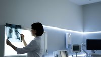 Female Asian doctor examining chest X-ray film on illuminated medical light box in modern hospital radiology department for diagnostic healthcare analysis.