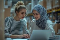 Young Caucasian woman and Middle Eastern woman in hijab working together on laptop in warehouse inventory management setting.