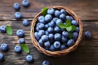 Fresh ripe blueberries in woven basket with green mint leaves on rustic wooden background, scattered berries around, macro food photography.