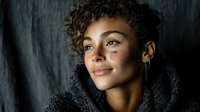 Young mixed-race woman with curly hair and facial bruises wearing dark hoodie against textured wall background for domestic violence awareness campaigns.