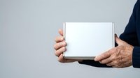 Senior man with light skin tone holding blank white tablet device with both hands against neutral gray background for product display or mockup purposes.