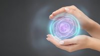 Female hands protecting glowing purple and blue energy sphere with swirling light effects on gray background for wellness and spiritual concepts.