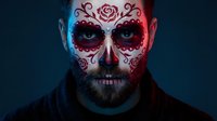Day of the Dead sugar skull face paint on bearded man with intricate red floral designs and decorative patterns for Halloween celebration and cultural events.