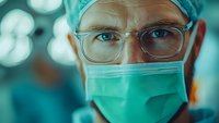 Close up portrait of medical professional wearing surgical mask, cap and glasses in operating room with dramatic lighting and teal tones in background.