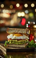 Gourmet avocado toast sandwich with fried egg on artisan bread served on wooden board in cozy restaurant with warm bokeh lights background.