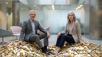 Successful mature business partners sitting on pile of golden coins in modern office lobby celebrating financial achievement and wealth growth.