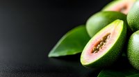 Fresh cut green papaya with pink flesh and black seeds on dark background with green leaves, macro food photography showing tropical fruit texture and details.