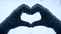 Hands in gray knitted gloves forming a heart shape against a snowy winter background, symbolizing love and warmth in cold weather.