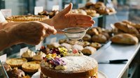 Professional baker dusting powdered sugar on decorated cake in commercial bakery kitchen with fresh baked goods display for culinary business operations.