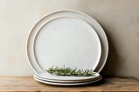 Rustic white ceramic plates stacked on wooden surface with fresh rosemary sprig, neutral background, minimalist kitchen styling.