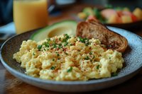 Creamy scrambled eggs garnished with fresh herbs and spices on gray ceramic plate, served with toasted bread and orange juice for breakfast meal.
