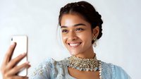 Young South Asian woman in traditional light blue embroidered dress and ornate gold necklace taking selfie with smartphone, showing bright genuine smile.