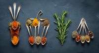 Aromatic spices and herbs arranged on vintage spoons with fresh rosemary on dark slate background for culinary cooking and seasoning recipes.