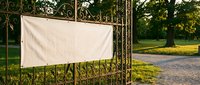 White blank banner hanging on ornate metal fence gate in park setting with trees and pathway for advertising mockup display.