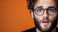 Young bearded man with curly hair and glasses showing surprised expression against orange background, close up portrait with copy space.