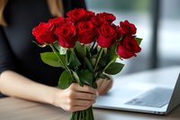 Woman holding fresh red roses bouquet while working on laptop at desk, romantic lifestyle moment captured in soft natural lighting.