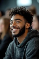 Happy young African American man with curly hair smiling in gray hoodie at social gathering or event with blurred people in background.