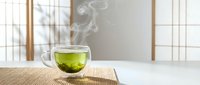 Steaming hot green tea in transparent glass cup on bamboo mat with traditional Japanese room interior background creating peaceful zen atmosphere.