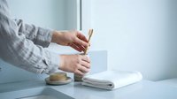 Professional woman writing handwritten note at clean white desk with coffee cup and notebook in bright modern office workspace for business communication.