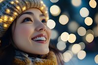 Young Asian woman in winter hat and scarf smiling while looking up at festive bokeh lights during holiday season, expressing joy and wonder against blurred background.
