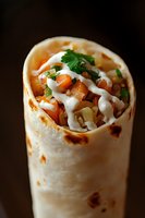 Fresh burrito filled with diced tomatoes, chopped herbs, and drizzled with creamy white sauce on dark background, close-up view of Mexican street food.