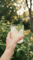 Hand holding glass of refreshing lemonade with fresh mint leaves and ice cubes outdoors in summer garden setting with blurred green foliage background.