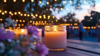 Glowing candle in glass jar on wooden deck at dusk with string lights and purple flowers creating romantic outdoor ambiance. Perfect for lifestyle blogs.