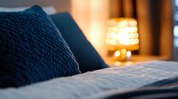 Cozy bedroom interior with navy blue pillow on white bedding, warm lamp glow in background creating intimate atmosphere for relaxation and comfort.
