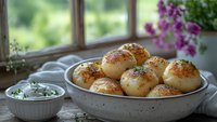 Freshly baked homemade dinner rolls with sesame seeds served in ceramic bowl, herb butter spread aside, purple flowers and window view background.