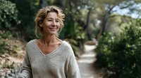 Happy mature blonde woman in cozy sweater smiling outdoors in natural park setting for lifestyle and wellness content.