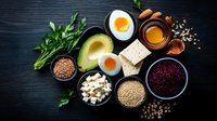 Healthy protein sources and superfoods arranged in bowls on dark wooden background, including avocado, eggs, tofu, grains, and fresh herbs.