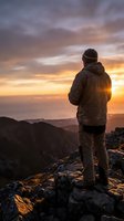 Man standing on rocky mountain peak watching golden sunset over valley landscape, wearing winter jacket and beanie, contemplating nature view for travel and adventure concepts.
