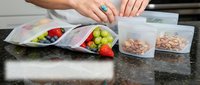 Woman's hands organizing meal prep containers with fresh fruits and cereals on dark granite countertop for weekly food planning and healthy eating.
