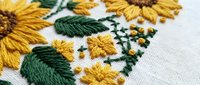 Hand embroidered sunflower pattern on white linen fabric with golden yellow petals and green leaves showcasing traditional needlework craftsmanship.