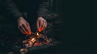 Hands lighting small campfire with glowing embers and flames in dark outdoor setting. Survival skills and wilderness camping concept.