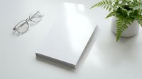 Clean white workspace with blank book, eyeglasses and green fern plant on marble surface for business planning, reading or creative writing projects.