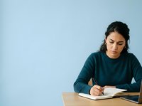 Woman with dark hair and olive complexion, wearing dark teal long-sleeve shirt, writing in notebook at wooden desk against light blue background. Suitable for education, business, and professional content.