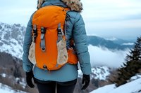 Female hiker wearing winter jacket and orange backpack standing on snowy mountain peak with foggy valley and mountain ranges in background.
