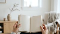 Person with light skin tone holding open blank notebook in cozy living room with natural light, suitable for lifestyle, education, journaling, and wellness content.
