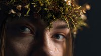 Close-up portrait of young Caucasian woman with blue eyes wearing natural forest crown made of moss, twigs, and small lights against dark background.