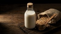 Fresh soy milk in glass bottle with cork stopper and raw soybeans spilling from burlap sack on rustic wooden table for healthy plant-based nutrition.