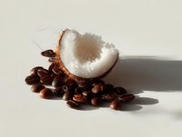 Fresh coconut slice resting on scattered coffee beans against neutral background, creating contrast between tropical and caffeinated ingredients.