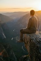 Young man sitting on cliff edge overlooking mountain valley at golden hour sunrise, contemplating nature landscape for mindfulness and adventure concepts.